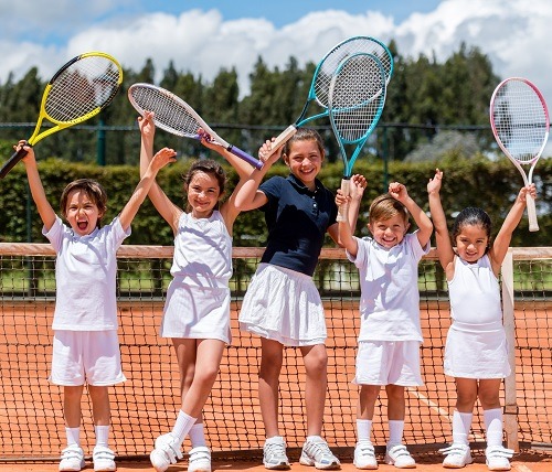 Mehrere lachende Kinder stehen mit erhobenen Tennisschlägern am Netz und feiern gemeinsam auf dem Platz.